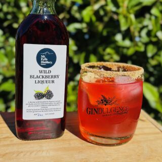 A Hedgerow Heist Cocktail in a glass next to a bottle of blackberry liqueur.